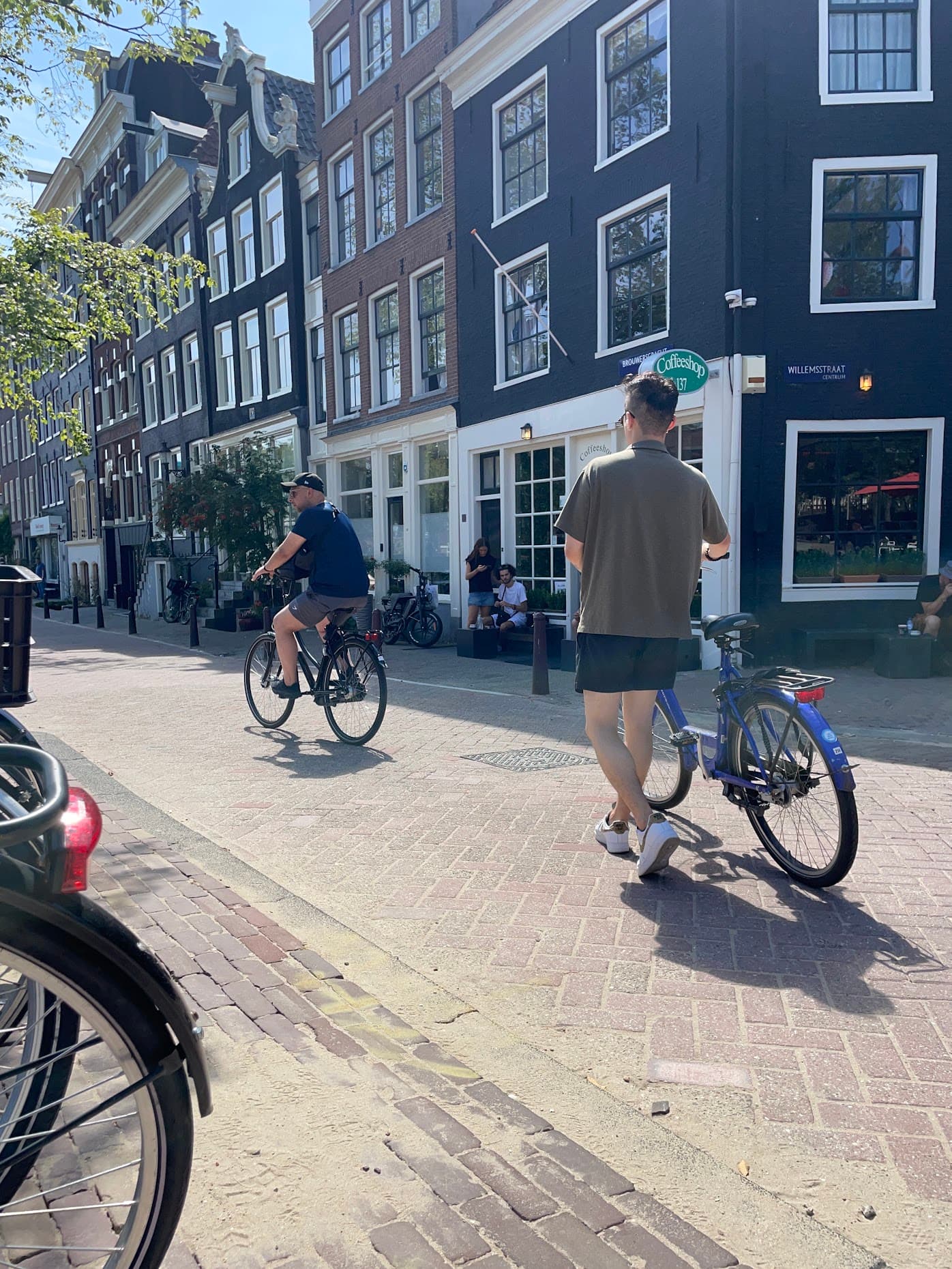 Cyclists passing a corner coffeeshop on a brick street lined with dark canal houses in Amsterdam